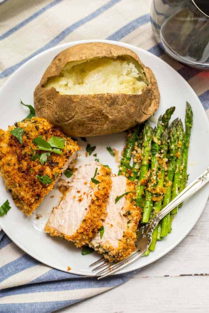 A white dinner plate with a baked potato, asparagus and crunchy baked pork chop that's sliced