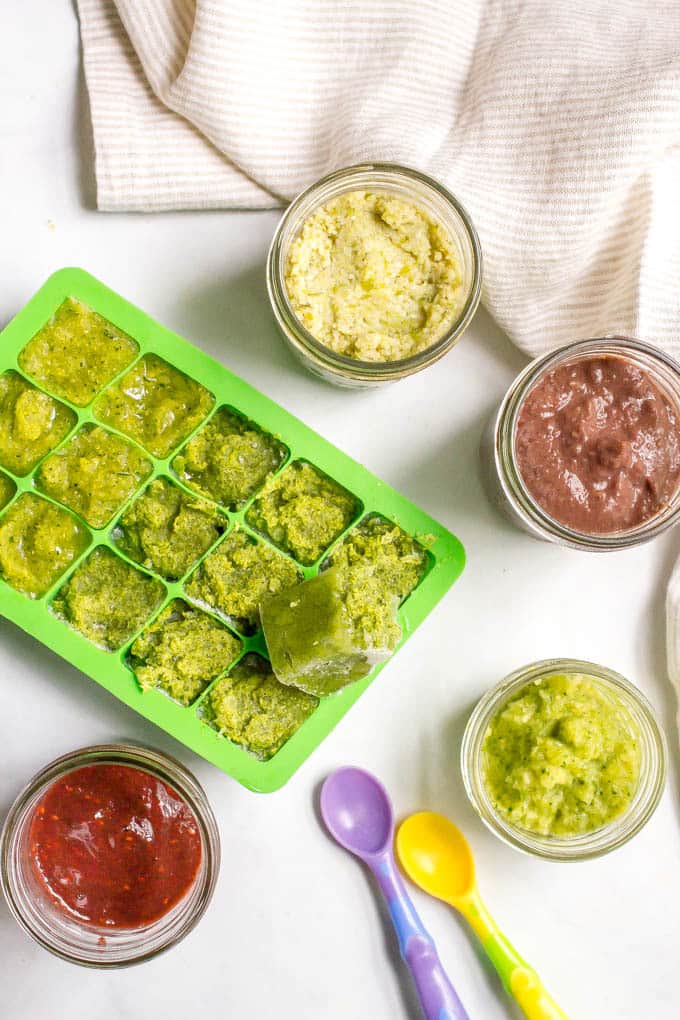 An array of different homemade baby foods in small glass jars next to a green silicone mini freezer cube tray also filled with a homemade baby food puree.
