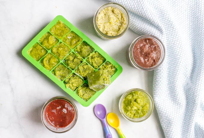 Small glass jars of different homemade baby food mixtures and a green silicone freezer tray with a green baby food puree in it.
