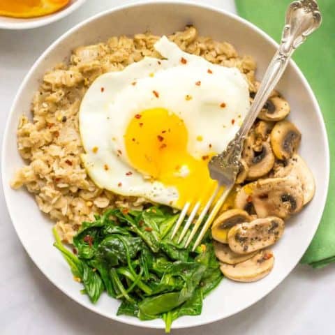 Savory oatmeal bowl with sauteed mushrooms, spinach and a fried egg on top in a bowl overhead shot with fork in runny egg yolk