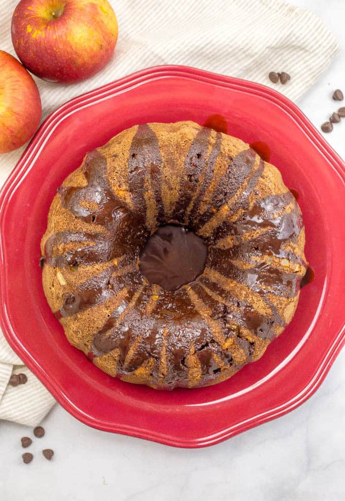Chocolate sauce topped apple cake with chocolate chips on a round red platter with apples and chocolate chips to the side.