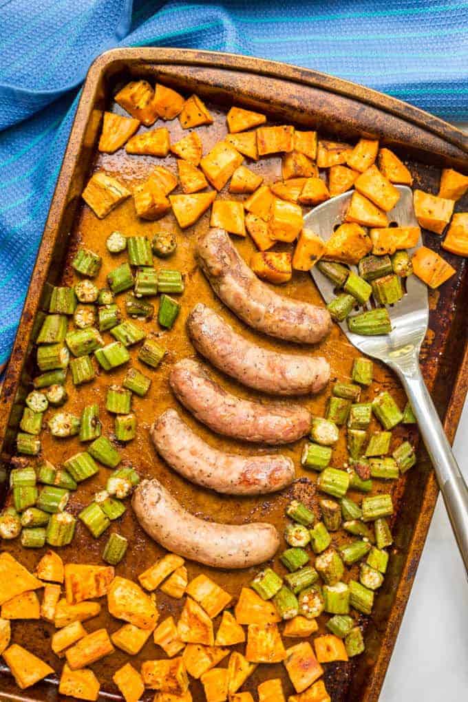 A sheet pan with roasted sweet potatoes and okra and a row of roasted turkey sausages in the middle and a spatula resting on the pan.