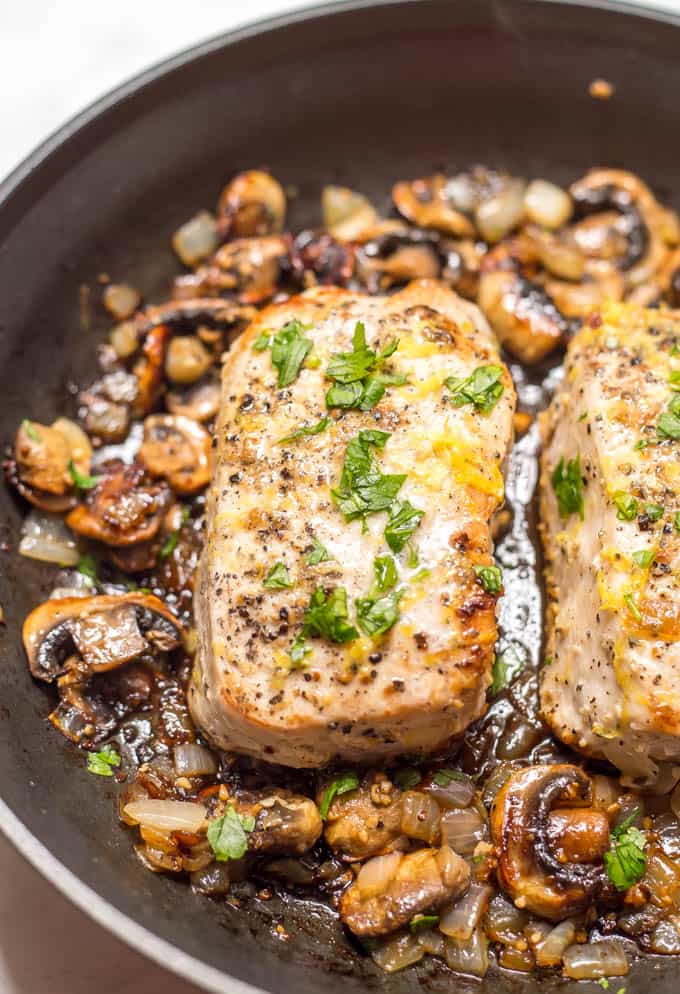 Close up of a pork chop being seared in a skillet with sautéed onions and mushrooms to the side.