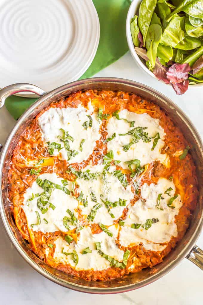 A large all clad skillet with a lasagna mixture topped with fresh mozzarella cheese and julienned basil.