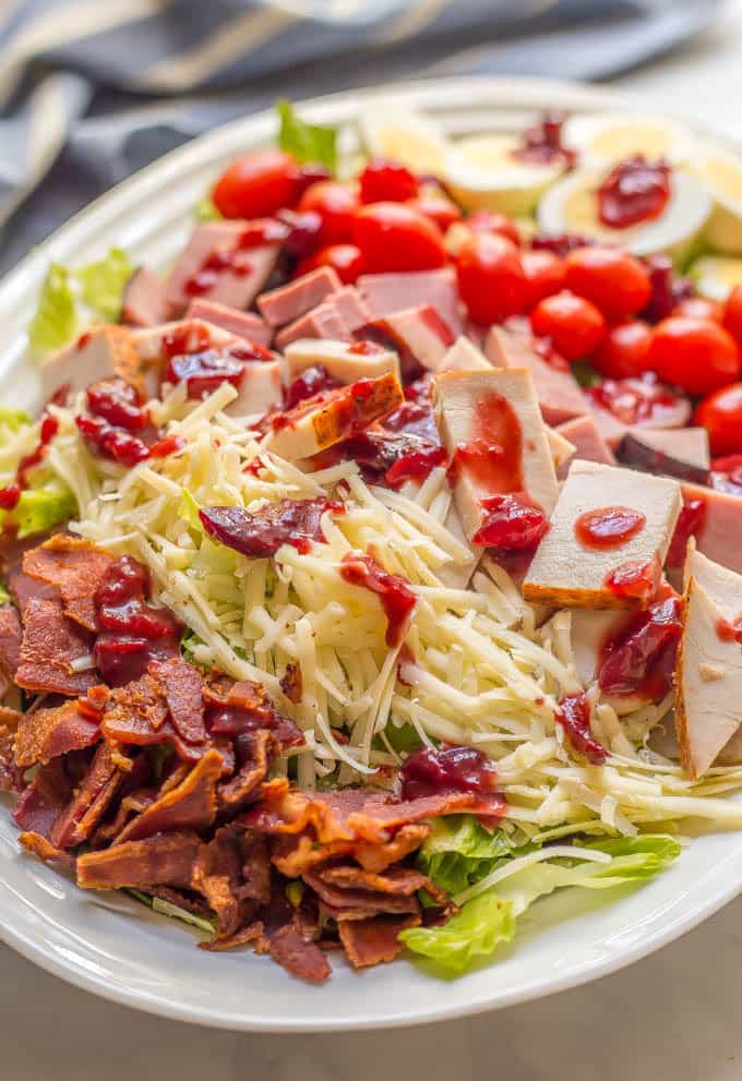 Close up of an arranged chef salad with leftover Thanksgiving turkey and ham and rows of cheese, bacon, tomatoes and hard boiled eggs with a cranberry sauce dressing on top.