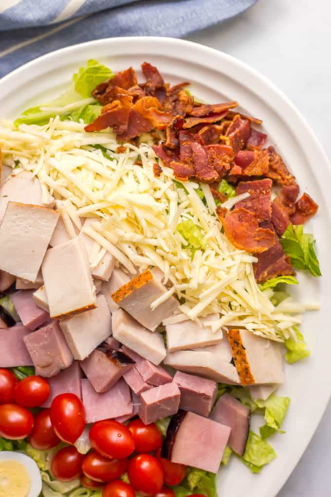 Close up of ingredients layered on a Thanksgiving chef salad in a white bowl: crumbled bacon, shredded cheese, turkey, ham and cherry tomatoes