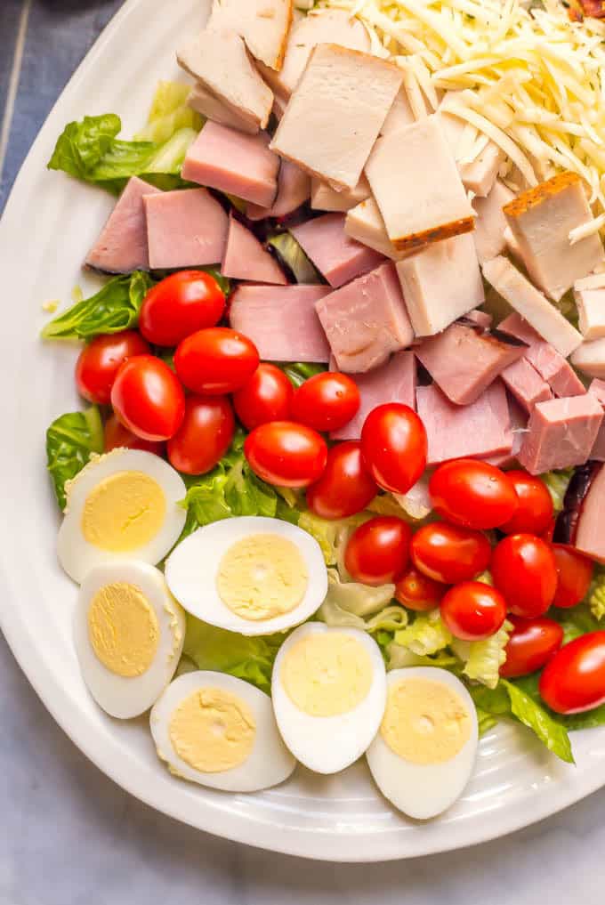 Close up of ingredients layered on a salad: hard boiled eggs, cherry tomatoes, ham, turkey and shredded cheese