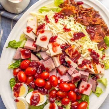 An arranged chef salad on a white platter with leftover Thanksgiving turkey and ham and rows of cheese, bacon, tomatoes and hard boiled eggs with a cranberry sauce dressing on top.