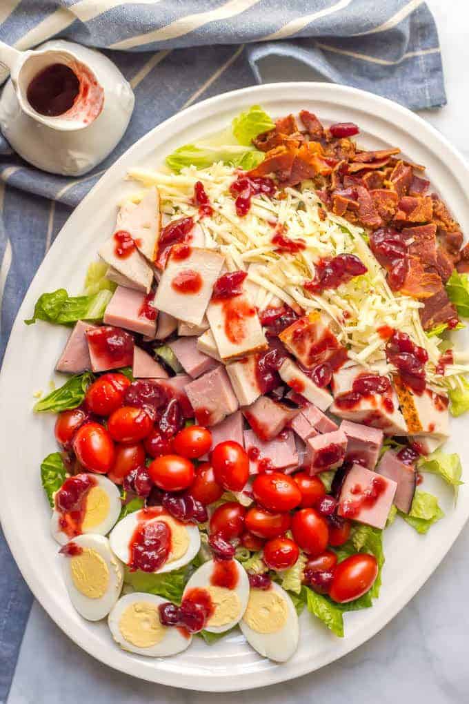 An arranged chef salad on a white platter with leftover Thanksgiving turkey and ham and rows of cheese, bacon, tomatoes and hard boiled eggs with a cranberry sauce dressing on top.