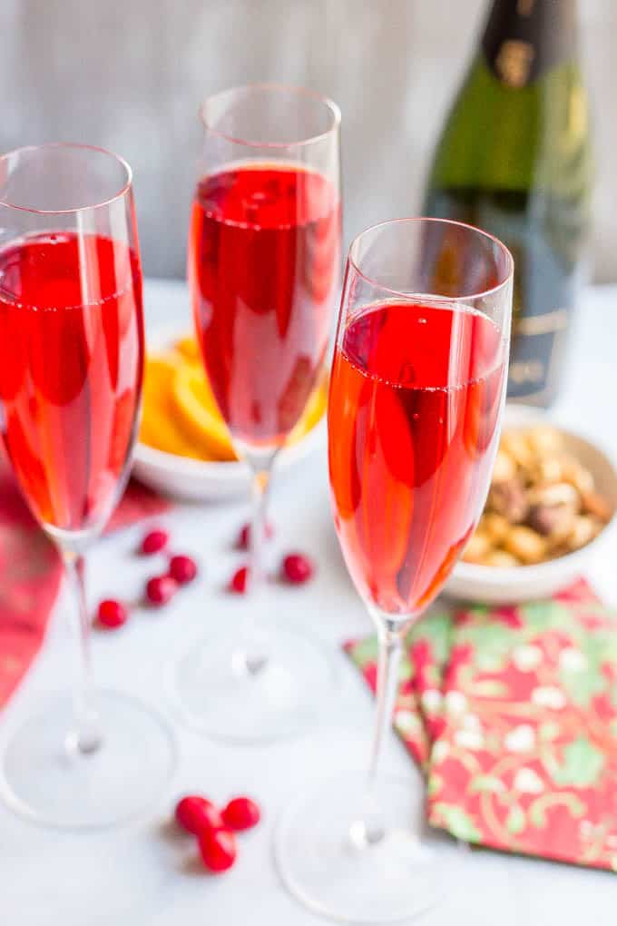 Vertical photo of 3 glasses filled with poinsettia cocktails with a bottle of chapmagne, orange slices and mixed nuts in the background