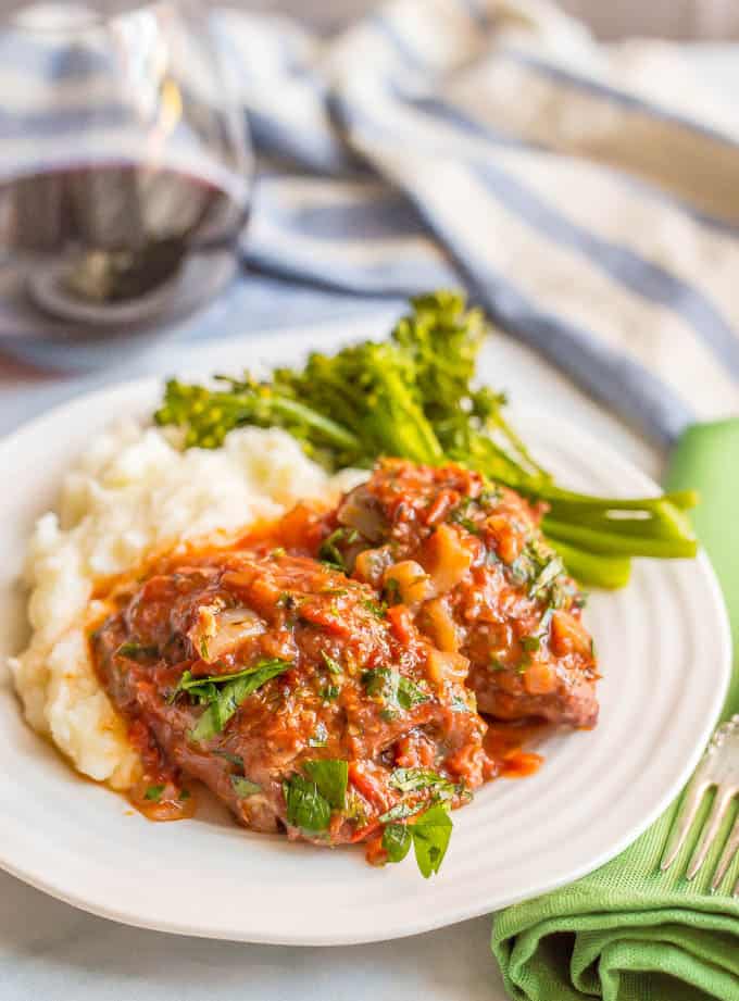 Slow cooker bistro chicken thighs served over mashed potatoes with broccolini and a glass of red wine in the background