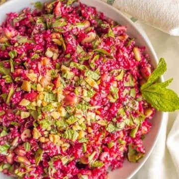 A chopped beet and quinoa salad in a white bowl with spinach, edamame and walnuts with a sprig of fresh mint on the size.