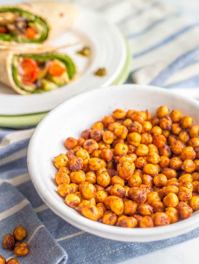 Crunchy BBQ roasted chickpeas in a white bowl with a blue striped towel underneath and a veggie wrap on a plate in the background