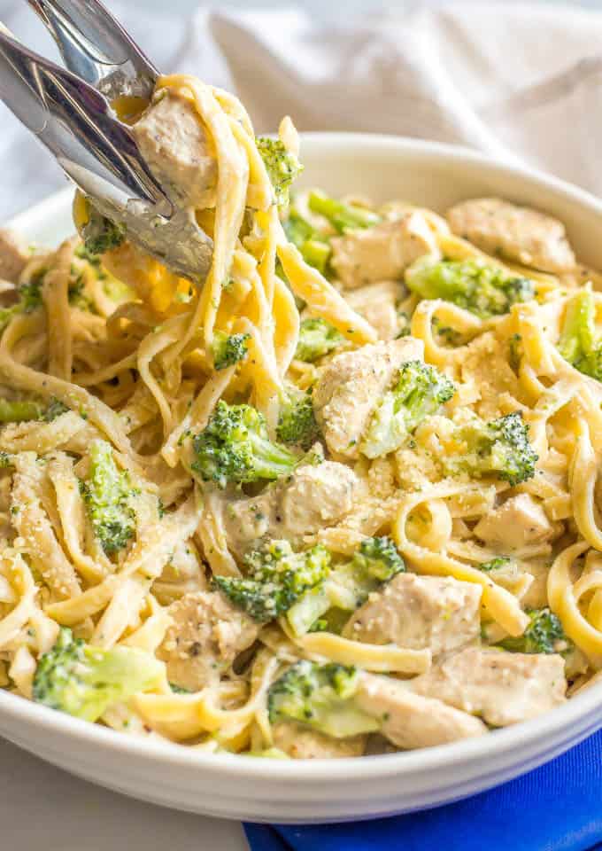 Healthy chicken Alfredo with broccoli in a large white serving dish with tongs taking out a scoop