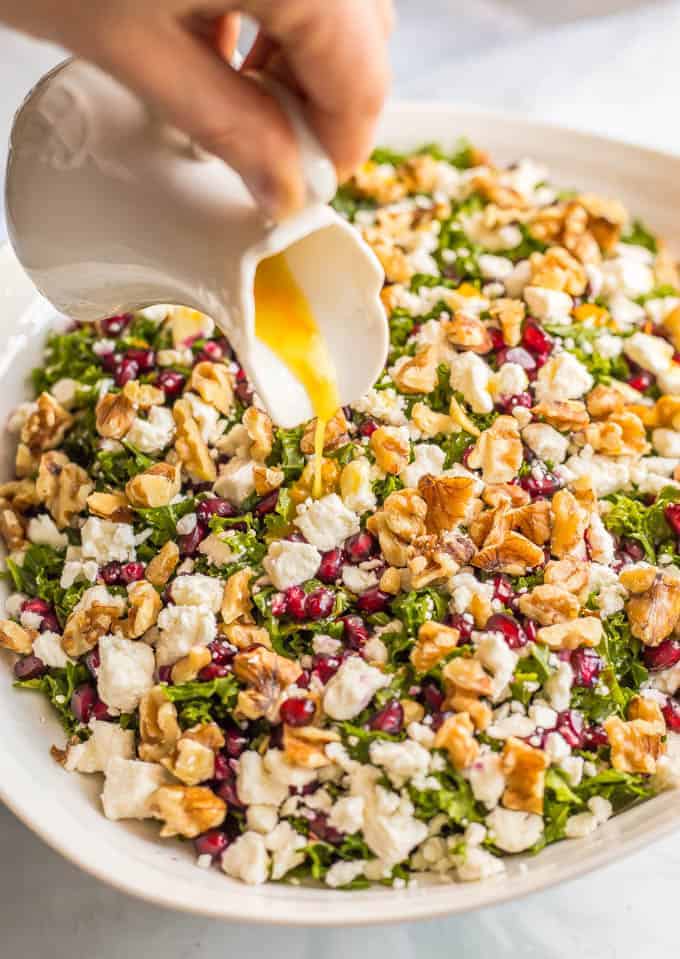 A hand pouring a citrus vinaigrette over a bowl of a colorful winter kale salad with walnuts, feta cheese and pomegranate arils mixed in.