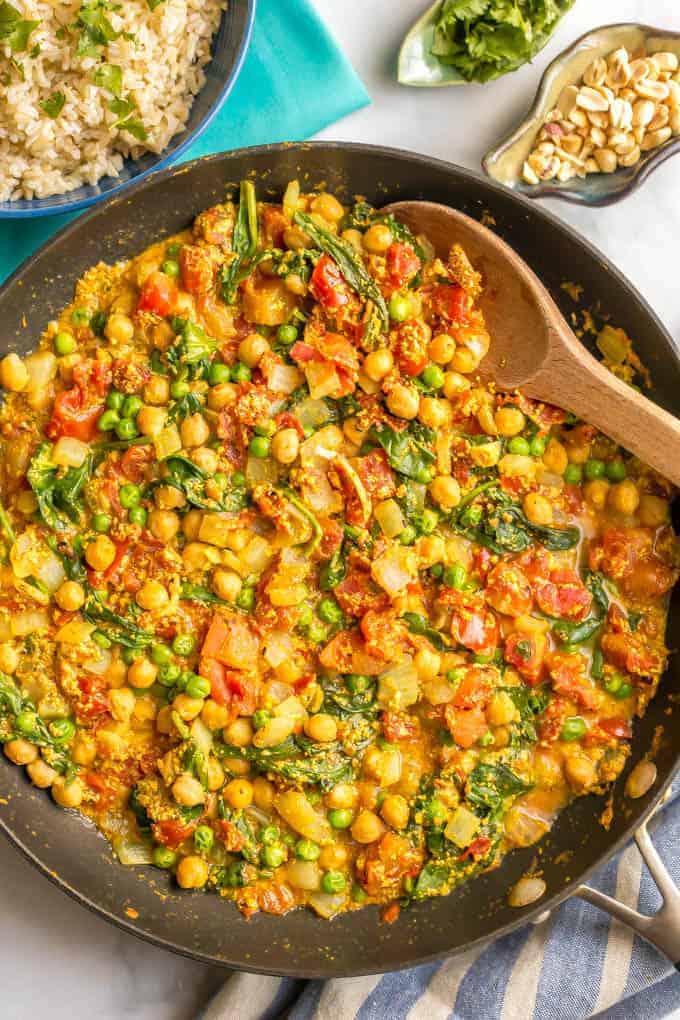 A large dark skillet with a vegetarian chickpea curry with spinach and tomatoes and a wooden spoon resting in the pan