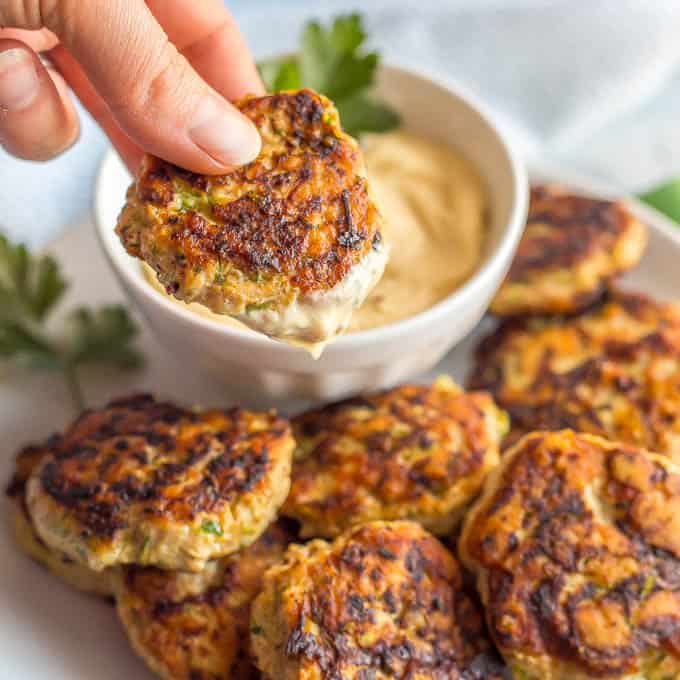 A single spicy chicken patty dipped into a creamy mustard sauce and being held up with a plate of chicken patties in the background