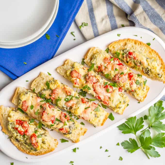 Slices of spinach artichoke stuffed French bread on a white serving plate with roasted red peppers, Parmesan cheese and fresh parsley on top
