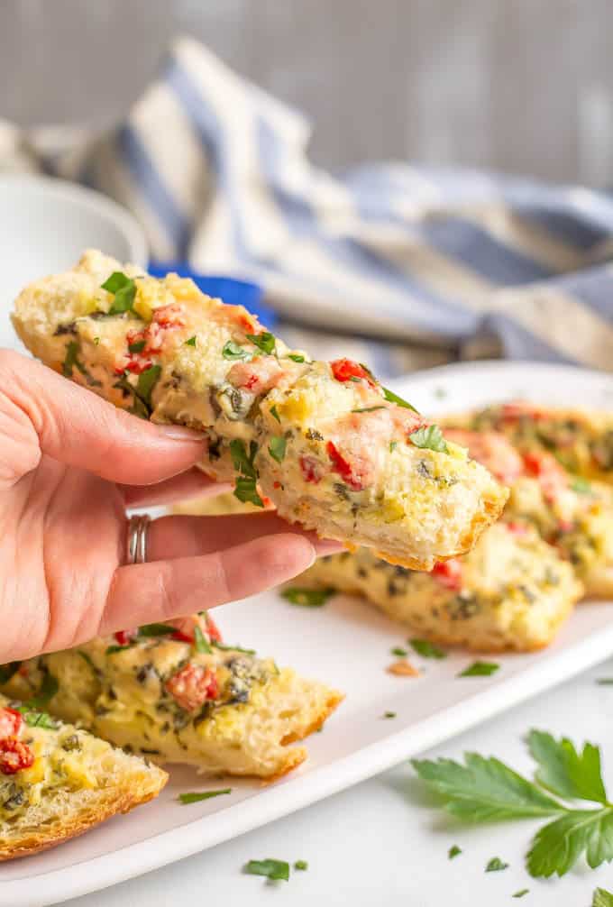 A hand holding a slice of baked French bread with spinach artichoke dip, roasted red peppers and fresh parsley