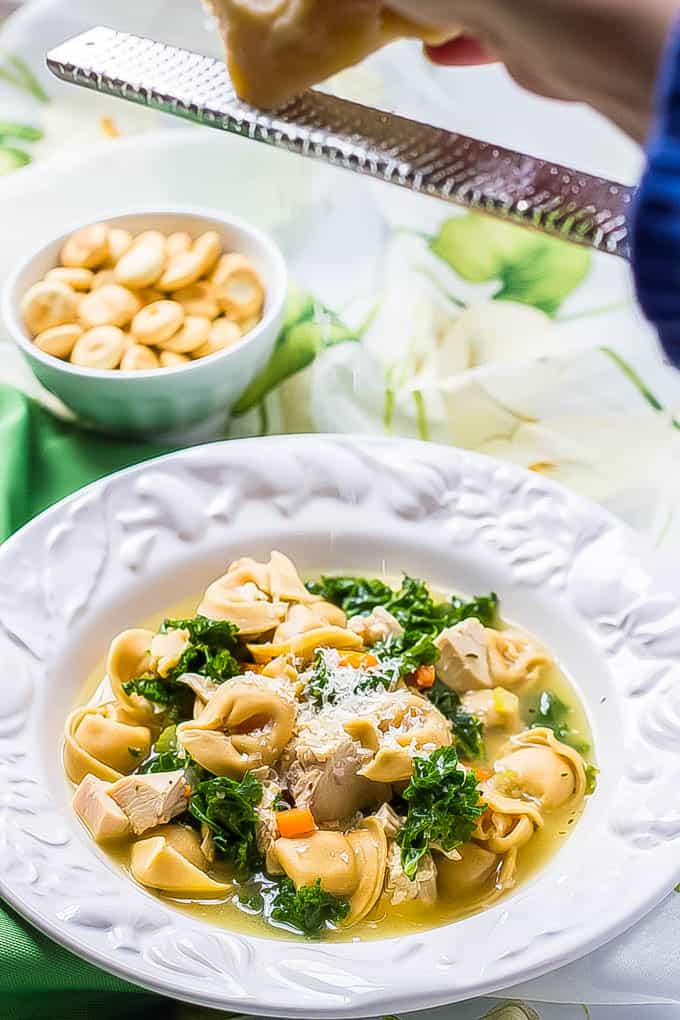 Parmesan cheese being grated over a bowl of tortellini soup with kale and chicken in a white bowl.