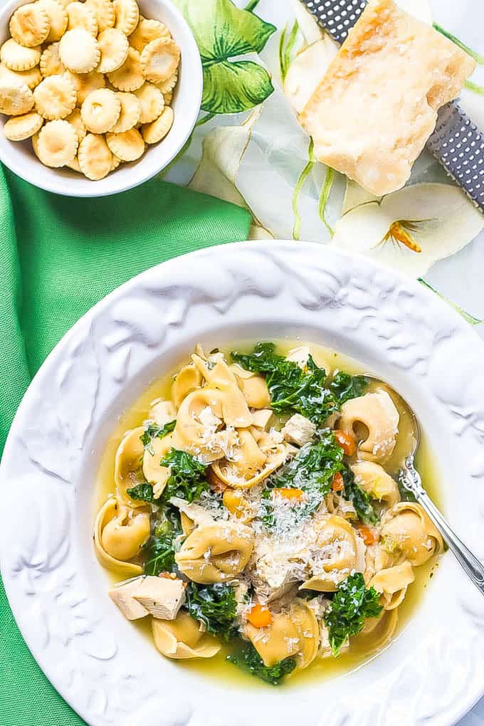 Overhead shot of chicken tortellini soup with kale being served in a white soup bowl with a spoon resting in the bowl and Parmesan cheese and oyster crackers nearby