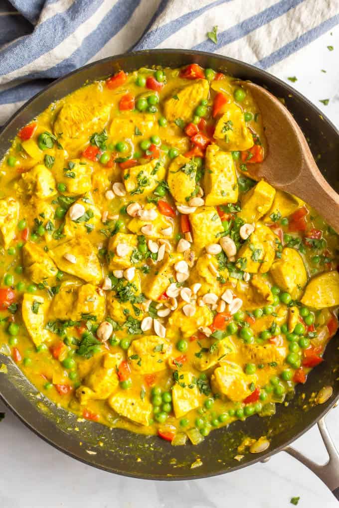 Overhead shot of one-pan coconut chicken curry in a large pan with peanuts and cilantro sprinkled on top and a wooden spoon lying in the pan