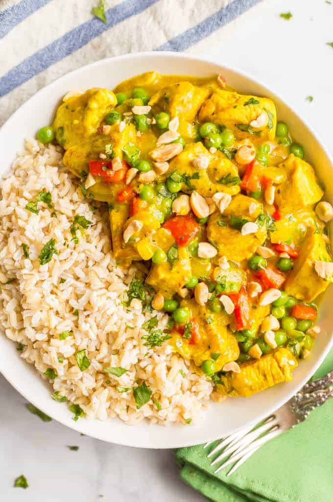 Overhead shot of coconut chicken curry in a white bowl beside steamed brown rice, with peanuts and cilantro sprinkled on top and forks lying nearby on a green napkin