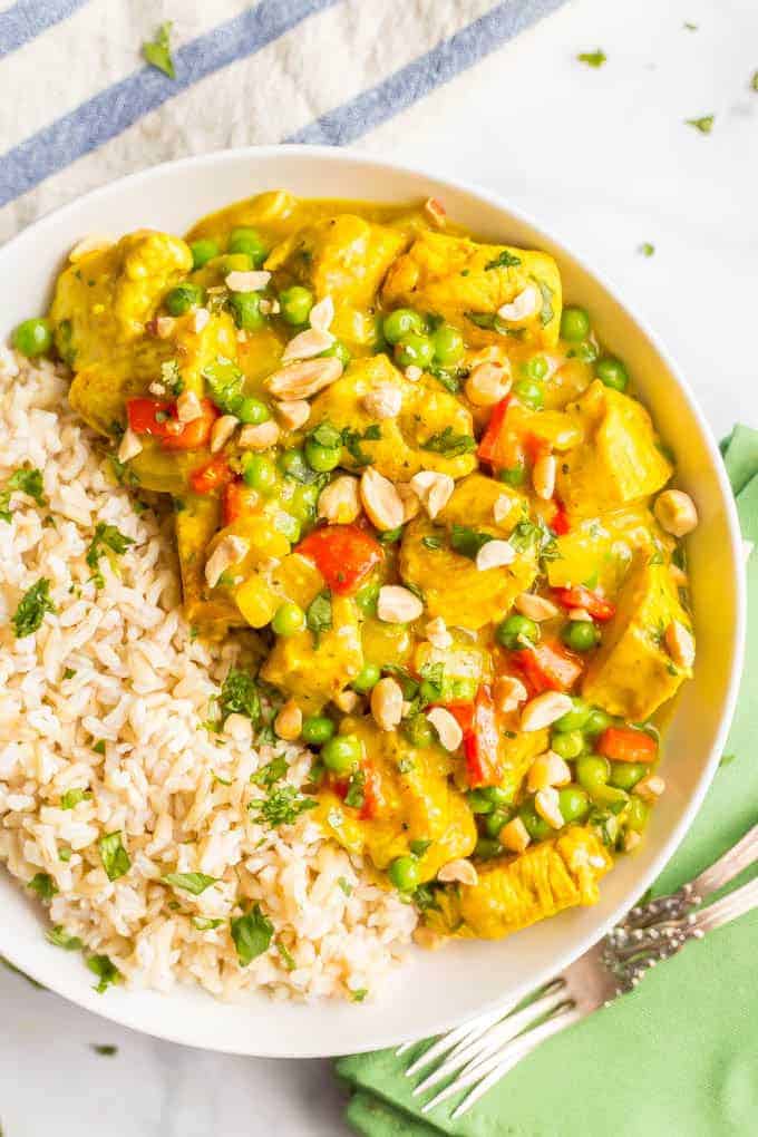 Close-up overhead shot of coconut chicken curry in a white bowl beside steamed brown rice, with peanuts and cilantro sprinkled on top and forks lying nearby on a green napkin