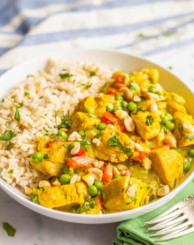 Close up side angle photo of coconut chicken curry in a white bowl beside steamed brown rice, with peanuts and cilantro sprinkled on top and forks lying nearby on a green napkin