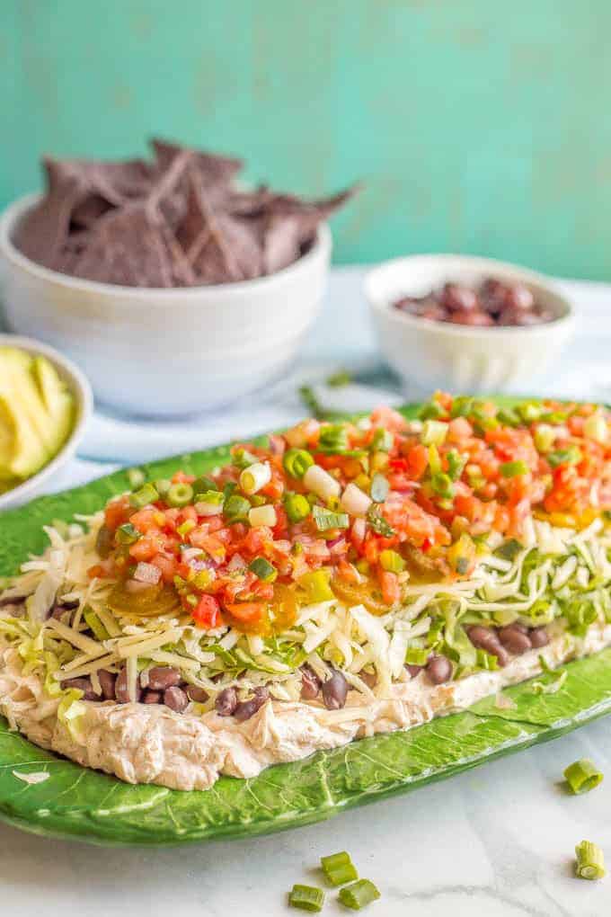 Side angle styled photo of healthy 7-layer Mexican dip on a green serving platter with bowls of chips, avocado and olives in the background and a sprinkling of green onions