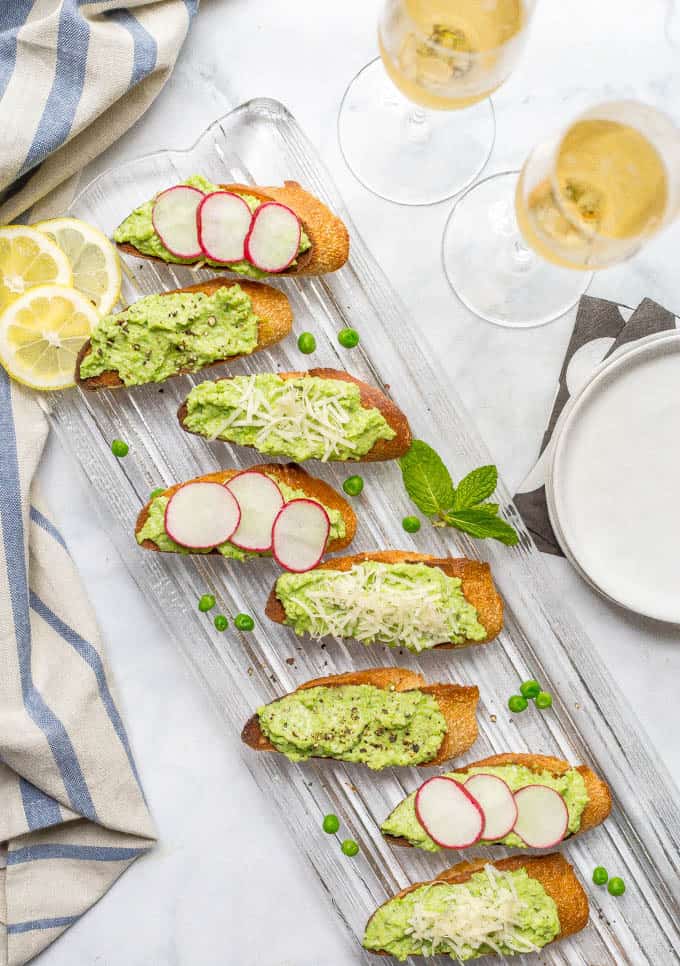 Overhead shot of baguette slices topped with healthy pea and ricotta dip, some with radish slices, some with Parmesan cheese and some with cracked black pepper and glasses of champagne nearby