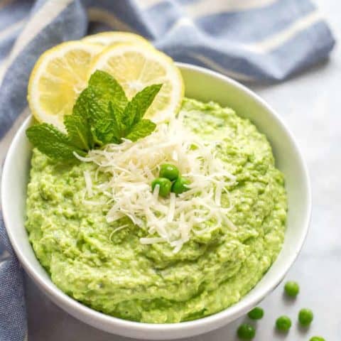 A bowl of healthy pea dip with Parmesan cheese sprinkled on top and a scattering of green peas nearby