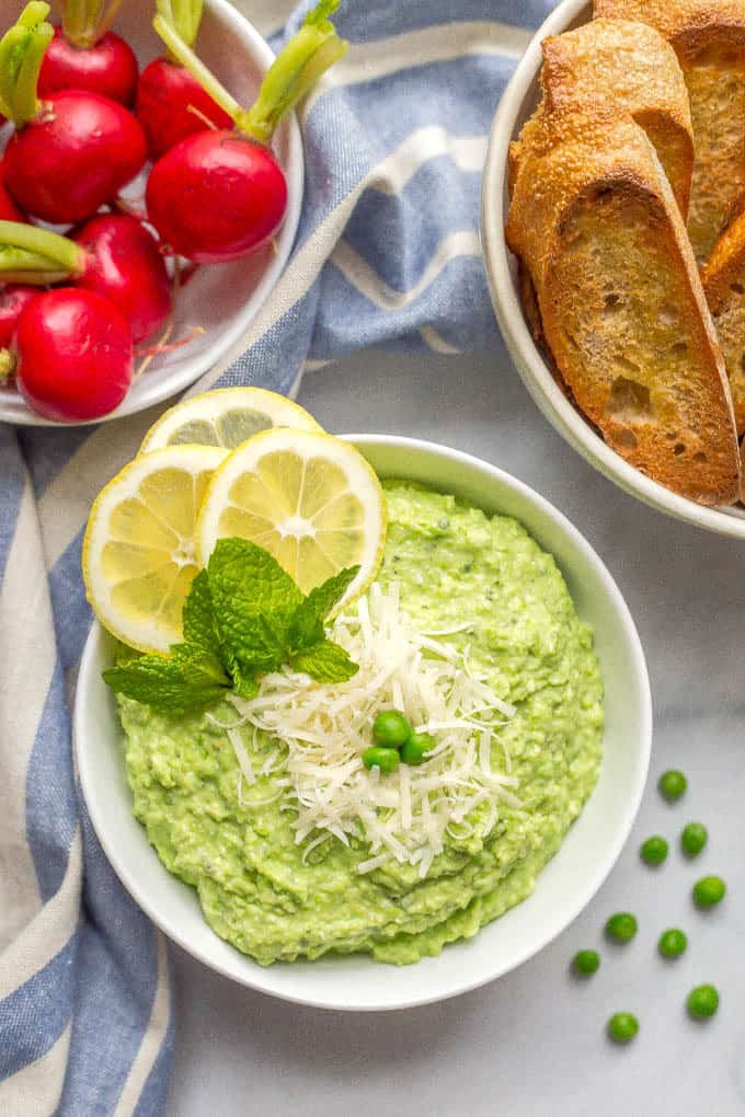 Overhead shot of a bowl of healthy pea dip with Parmesan cheese sprinkled on top and a scattering of green peas nearby and bowls of baguette slices and radishes in the background