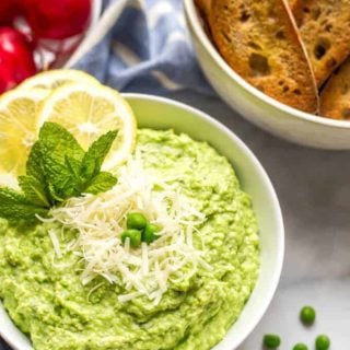 Side angle shot of a bowl of healthy pea dip with Parmesan cheese sprinkled on top and a scattering of green peas nearby and bowls of baguette slices and radishes in the background
