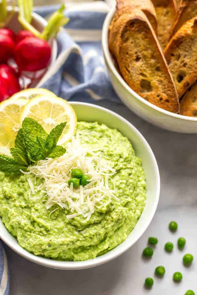 Side angle shot of a bowl of healthy pea dip with Parmesan cheese sprinkled on top and a scattering of green peas nearby and bowls of baguette slices and radishes in the background