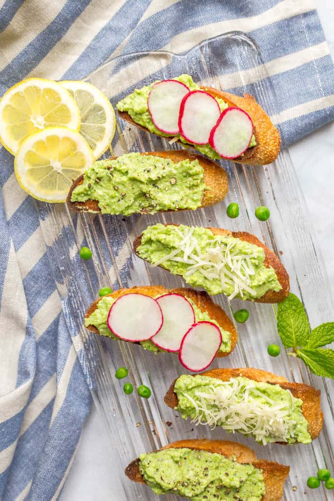 Overhead shot of baguette slices topped with healthy pea and ricotta dip, some with radish slices, some with Parmesan cheese and some with cracked black pepper