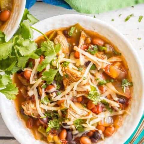 Overhead close-up shot of healthy slow cooker chicken chili served in a white bowl with shredded cheese and cilantro on top and a spoon on a turquoise napkin nearby