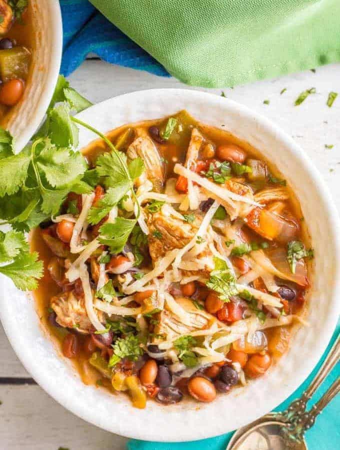 Overhead close-up shot of healthy slow cooker chicken chili served in a white bowl with shredded cheese and cilantro on top and a spoon on a turquoise napkin nearby.