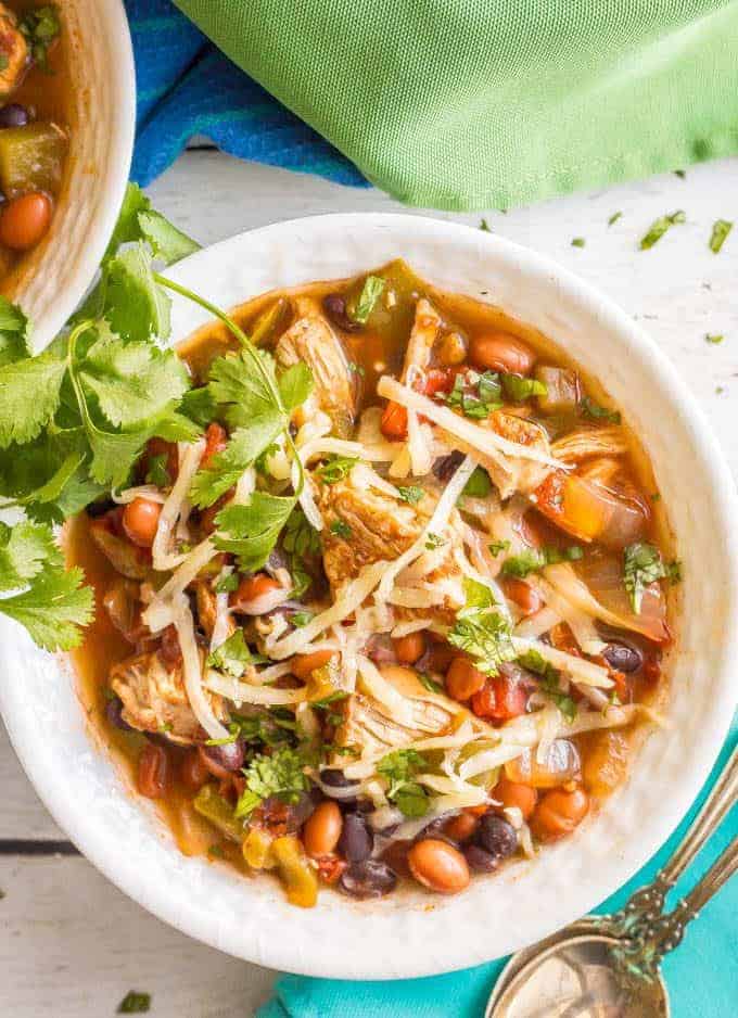Overhead close-up shot of healthy slow cooker chicken chili served in a white bowl with shredded cheese and cilantro on top and a spoon on a turquoise napkin nearby