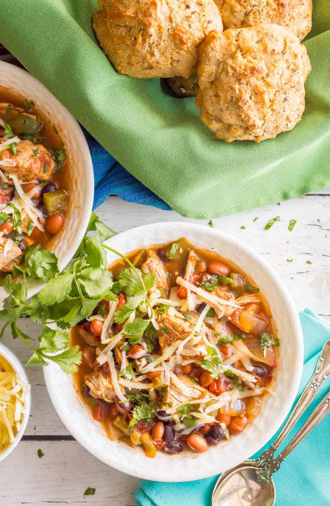 Overnhead shot of healthy slow cooker chicken chili served in a white bowl with shredded cheese and cilantro on top and biscuits in a bowl with a green napkin in the background