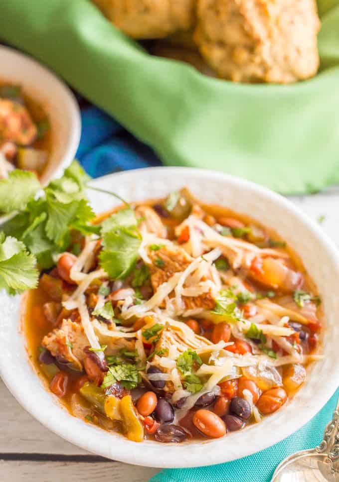 Healthy slow cooker chicken chili served in a white bowl with shredded cheese and cilantro on top and biscuits in a bowl with a green napkin in the background