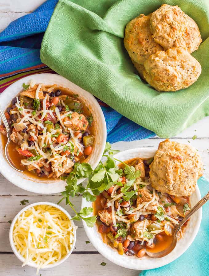 Healthy slow cooker chicken chili being served in two bowls with spoons in each and biscuits in a bowl with a green napkin nearby