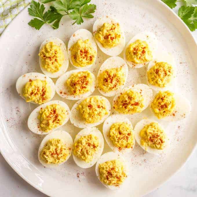 Square overhead photo of Mom's classic Southern deviled eggs sprinkled with paprika and served on a large, round white platter with parsley for a garnish