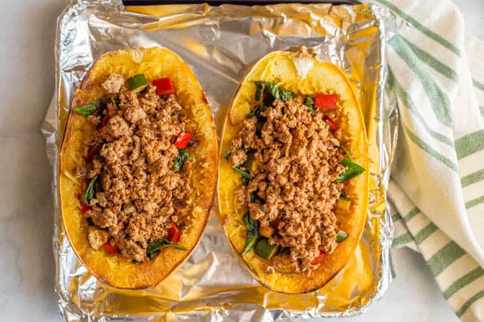 A roasted spaghetti squash cut in half and stuffed with veggies and ground turkey on a baking tray lined with foil and a green striped towel in the background