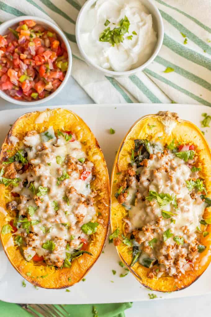 Turkey taco spaghetti squash boats with veggies and melted cheese being served on a large white plate with small bowls of pico de gallo and Greek yogurt nearby