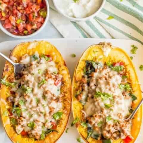Turkey taco spaghetti squash boats with veggies and melted cheese being served on a large white plate with forks stuck in each one and small bowls of pico de gallo and Greek yogurt nearby