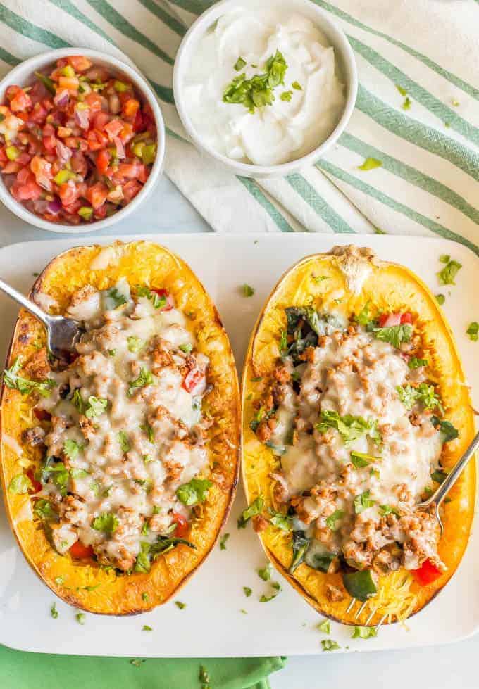 Turkey taco spaghetti squash boats with veggies and melted cheese being served on a large white plate with forks stuck in each one and small bowls of pico de gallo and Greek yogurt nearby