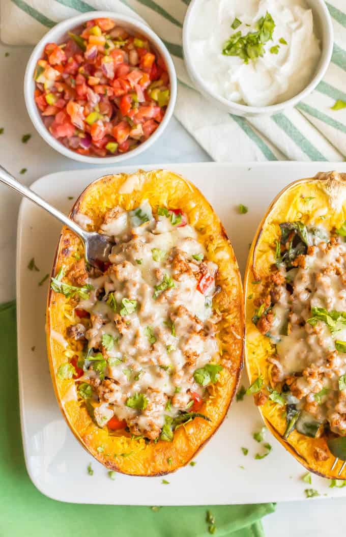A close-up of a turkey taco spaghetti squash boat with veggies and melted cheese being served on a large white plate with a fork stuck in and small bowls of pico de gallo and Greek yogurt nearby