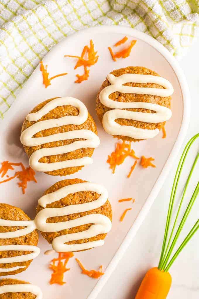 Overhead close-up photo of whole wheat carrot cake muffins with cream cheese frosting on a white plate with grated carrots and a whisk and green striped towel nearby