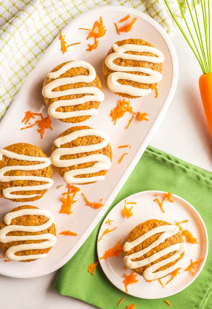 Overhead photo of several whole wheat carrot cake muffins with cream cheese frosting on a white plate with grated carrots and a separate individual plate, with a whisk and green striped towel nearby